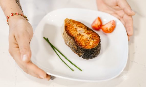 Close-up of grilled salmon served with tomatoes and chives on a plate, held by tattooed hands.